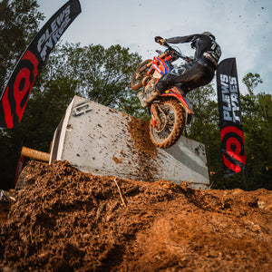 Enduro rider ascends a steep concrete obstacle on a muddy dirt bike during an off-road event, with Plews Tyres flags flanking the course and dirt flying into the air against a wooded backdrop.