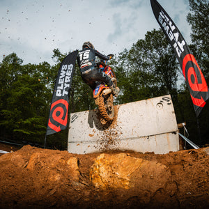 Motocross rider wearing jersey number 15 launches up a muddy vertical concrete wall obstacle during an off-road enduro race, with Plews Tyres flags on either side and dirt flying into the air against a forested background.