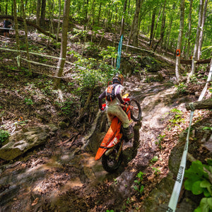 Dirt bike rider climbs a steep, muddy hill through a dense forest trail during an off-road race, surrounded by trees and race tape.