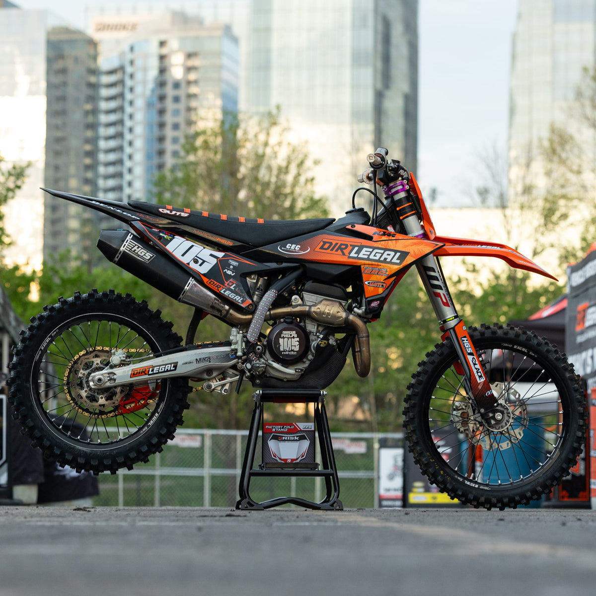 Orange and black dirt bike on a Risk Racing MX stand with cityscape in the background