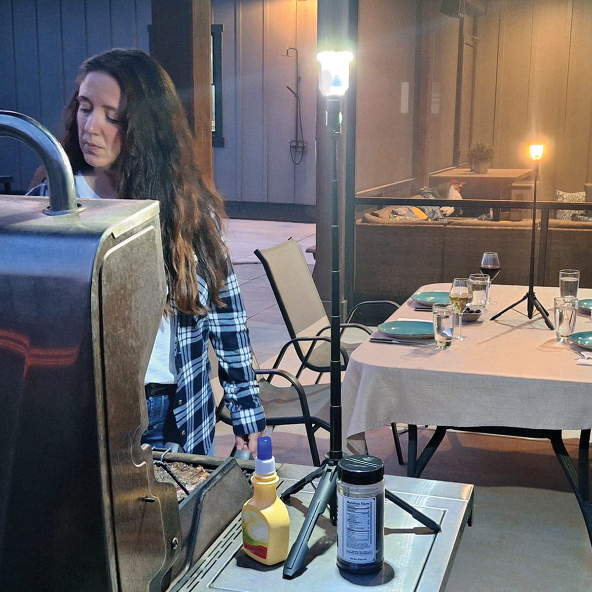 Woman grilling outdoors with a FLi-MORPHE lighting up here grilling experience. In the background, an outdoor table with place settings and wine glasses full. A FLi-MORPHE set to warm light sitting in the middle of the table.