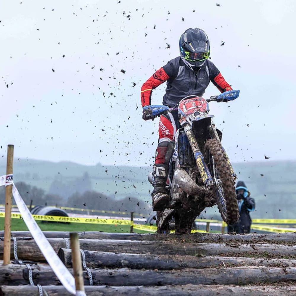 Motocross rider charges over a log obstacle on a muddy racecourse, flinging dirt into the air, during an off-road enduro event with rolling hills and spectators in the background.