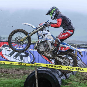 Endurocross racer launching over a row of tires on the track. He's wearing Risk Racing riding gear and his bike is equipped with Plews Tyres.