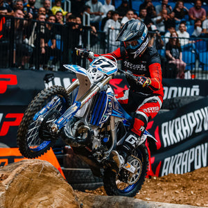 Endurocross Racer Launching up over some logs on the track. He's wearing Risk Racing riding gear and his bike is equipped with Plews Tyres. Stadium fans in the background.