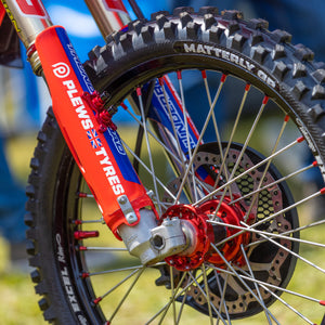 close up of a front tire and front forks of a dirt bike in an outdoor setting. The tire is a Plews Tyre Matterly GP and the fork covers are dawning a Plews Tyres sticker