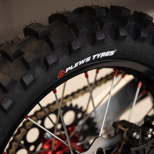Super close up on a rear dirt bike tire with the Plews Tyres logo prominently displayed in white against the black rubber.