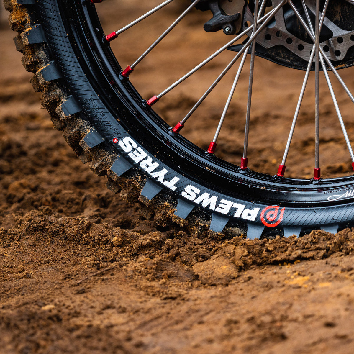 Super close up on a rear dirt bike tire with the Plews Tyres logo prominently displayed in white against the black rubber.