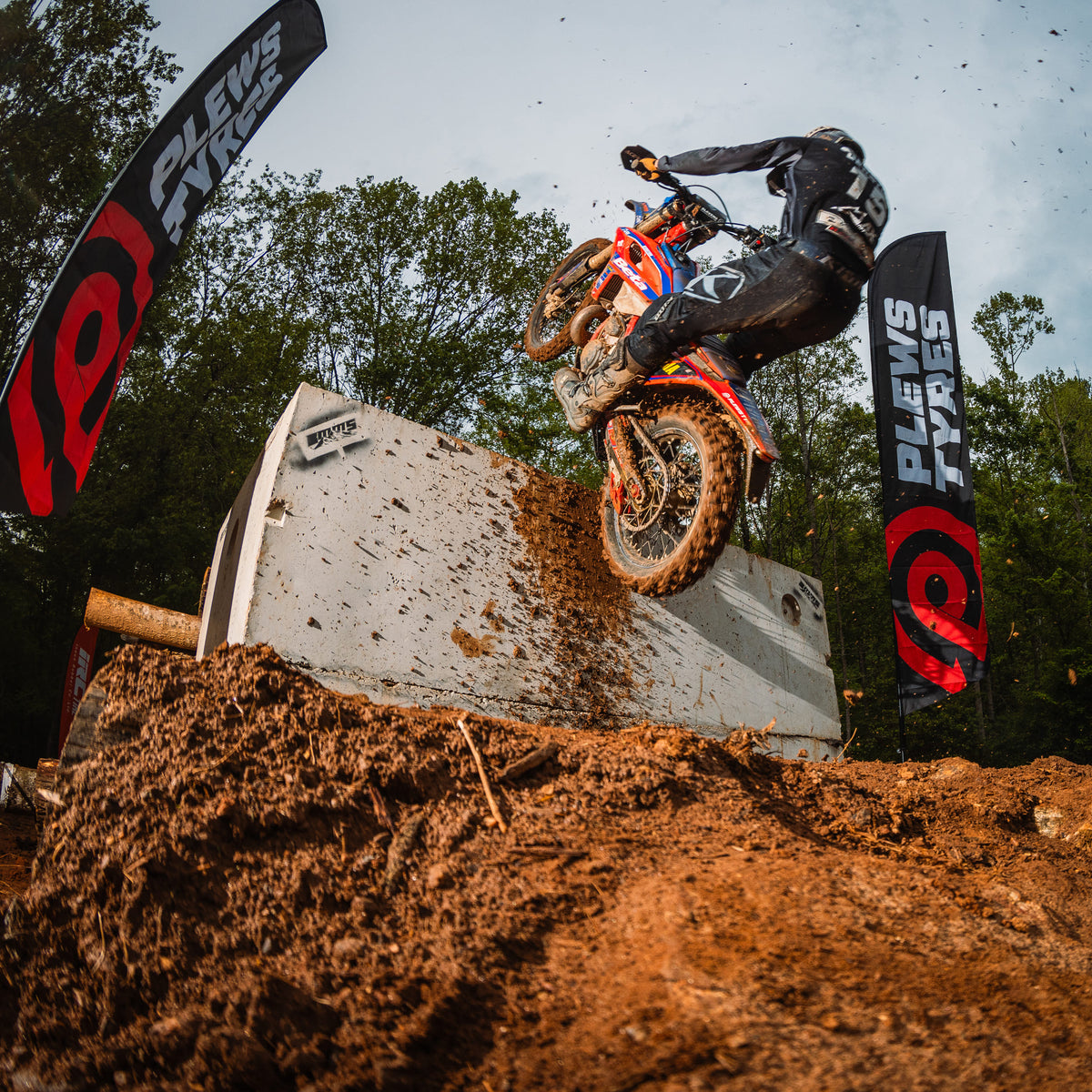 Enduro rider ascends a steep concrete obstacle on a muddy dirt bike during an off-road event, with Plews Tyres flags flanking the course and dirt flying into the air against a wooded backdrop.