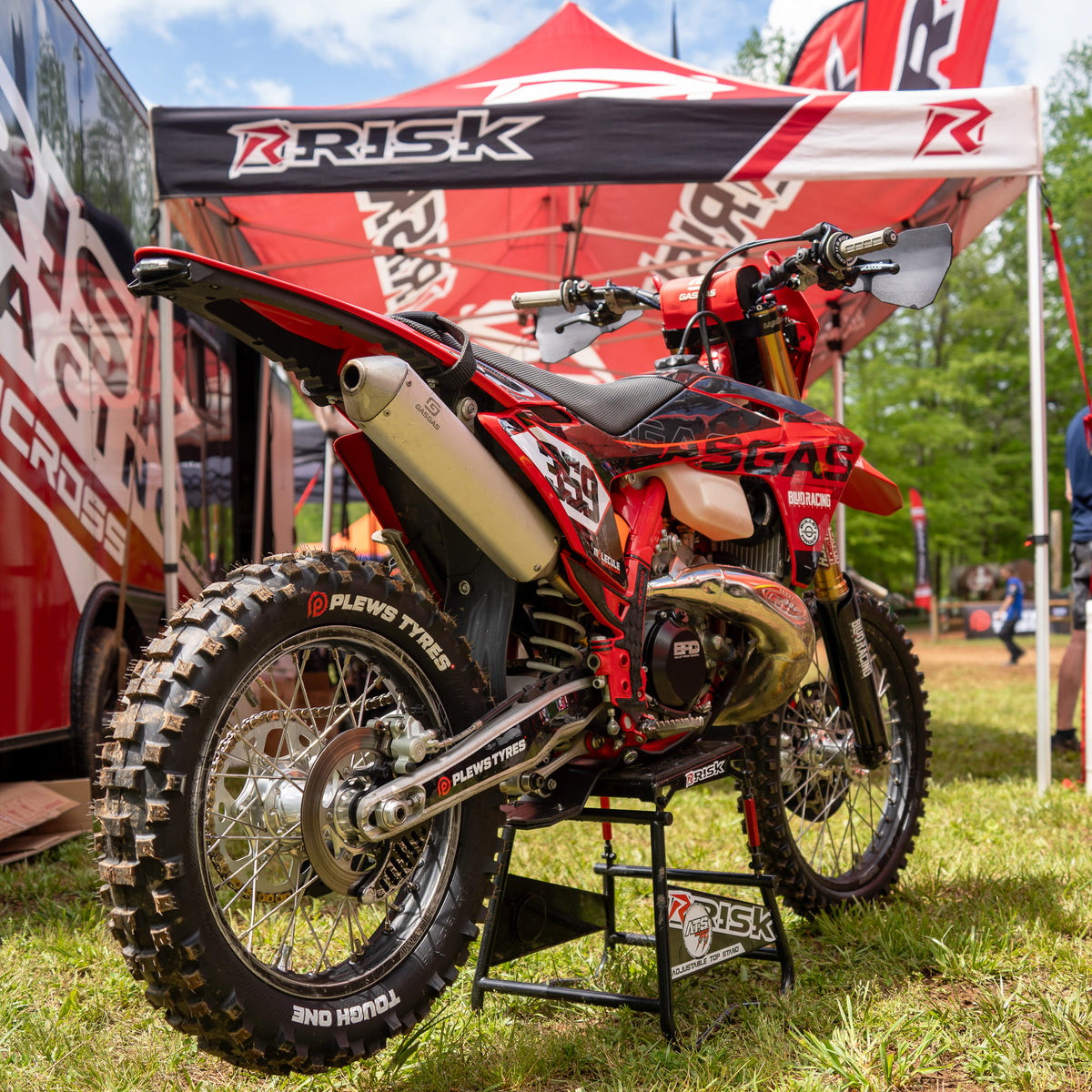 Red GASGAS motocross bike with Plews Tyres and Risk Racing branding displayed on a Risk Racing stand under a branded canopy tent at an outdoor racing event.
