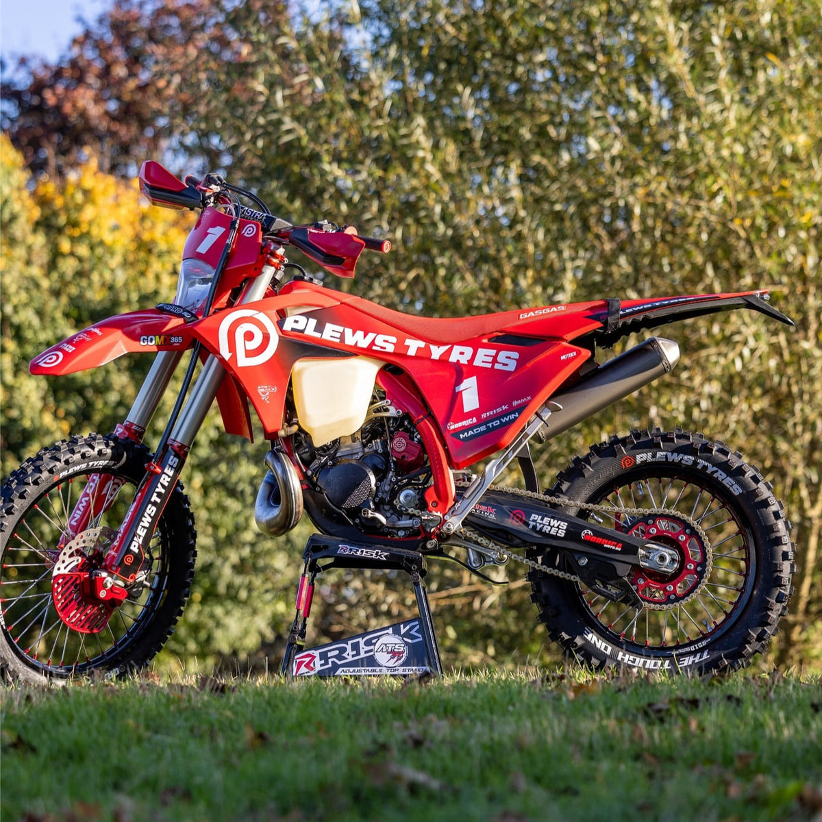 Red GASGAS dirt bike featuring Plews Tyres branding, displayed on a Risk Racing stand in a grassy outdoor setting with trees in the background.