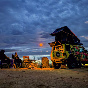 Overlanding camping scene featuring a male and female sitting in camping chairs near a jeep with roof tent. Everything is lit up by a FLi-PRO in amber mode.